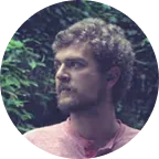 Man with curly hair and beard wearing a pink shirt looking to the side with green foliage in the background.