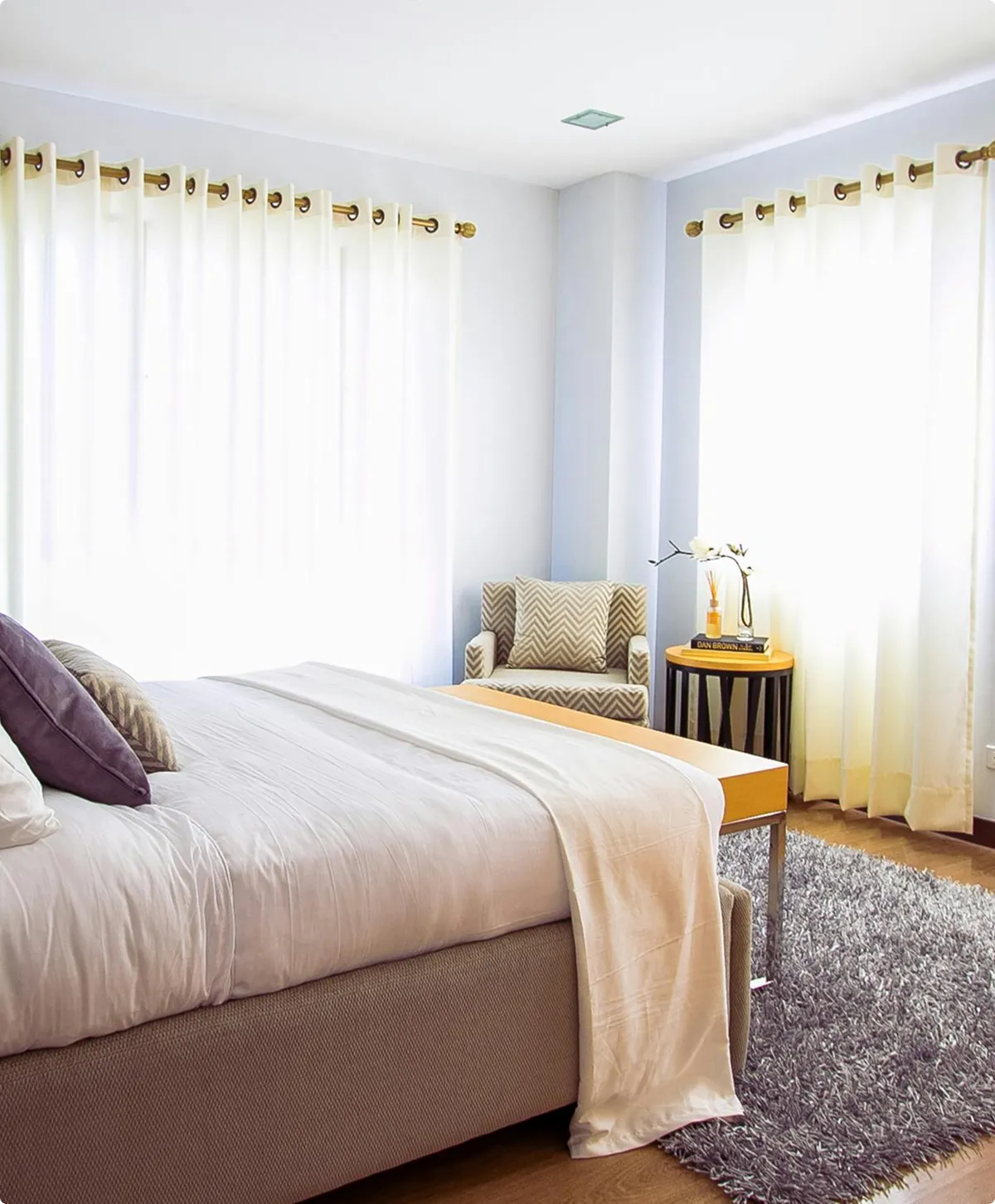 Bright bedroom with a beige upholstered bed, white bedding, purple and patterned pillows, a patterned armchair, round wooden side table with a book and flower, and a gray shag rug.