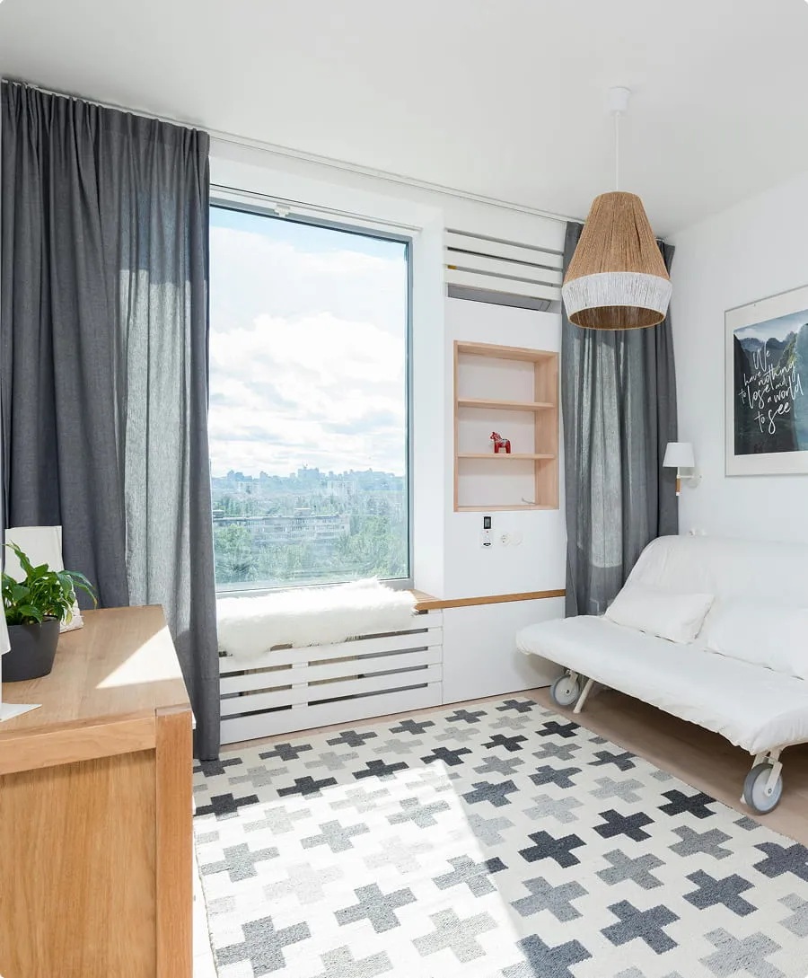 Bright modern living room with a large window, grey curtains, white futon on wheels, patterned rug, and wooden furniture.