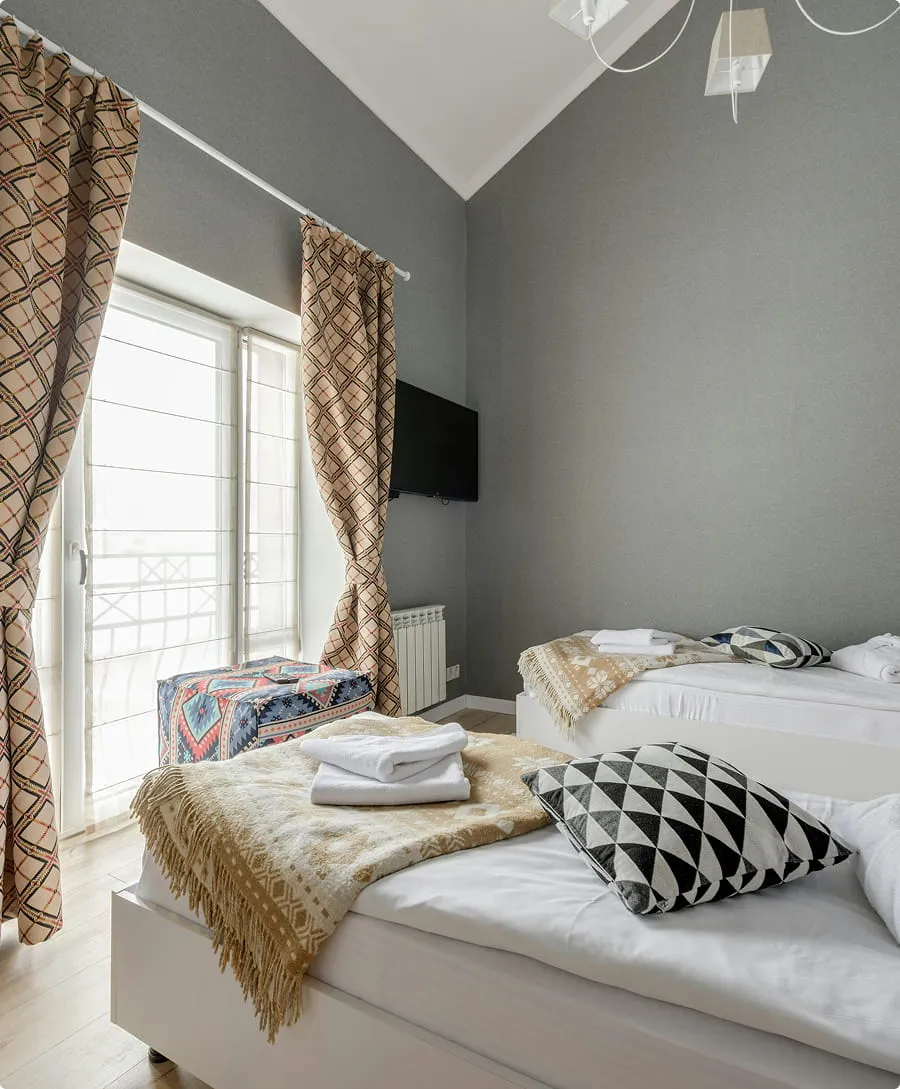 Minimalist bedroom with two single beds featuring patterned pillows and blankets, light wood flooring, patterned curtains, a wall-mounted TV, and a small colorful ottoman near large windows.