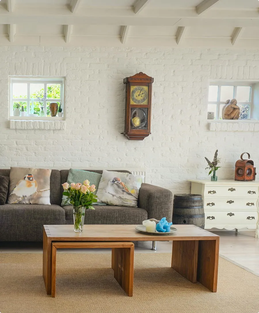 Cozy living room with a dark sofa featuring bird-patterned cushions, a wooden coffee table with flowers and candles, white brick walls, a vintage wall clock, and decorative items on a white dresser.