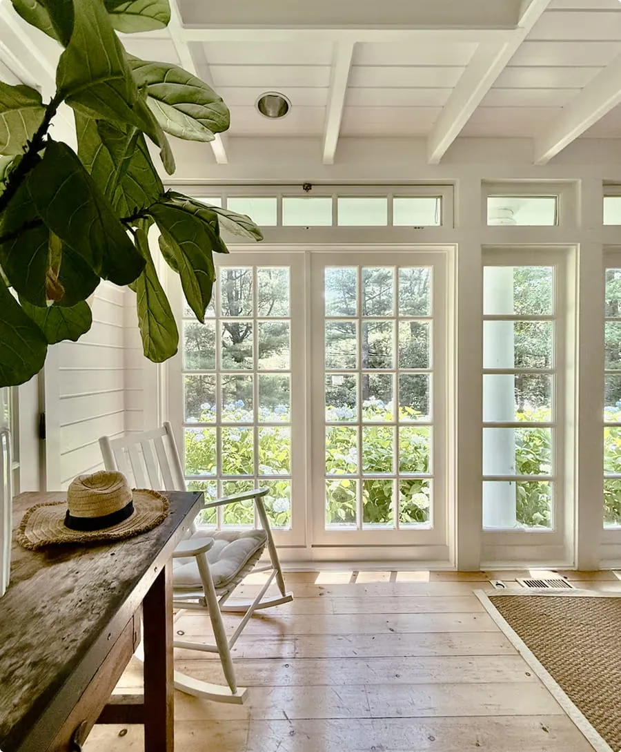 Bright room with wooden floor, white rocking chair, rustic table with a straw hat, and large windows showing green bushes and trees outside.