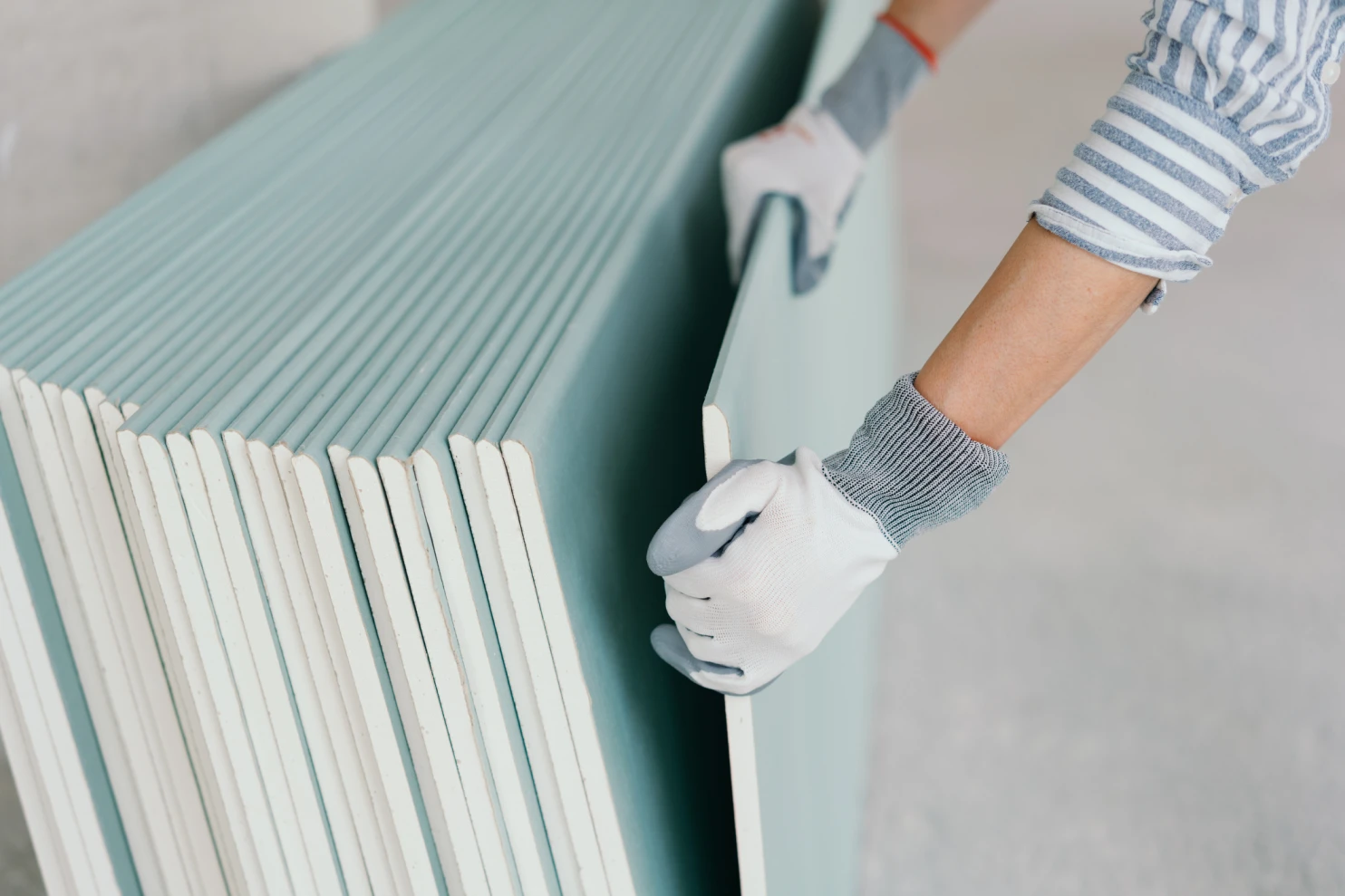 Eine Person mit Handschuhen hantiert im Innenbereich einer Baustelle mit gestapelten Gipskartonplatten.