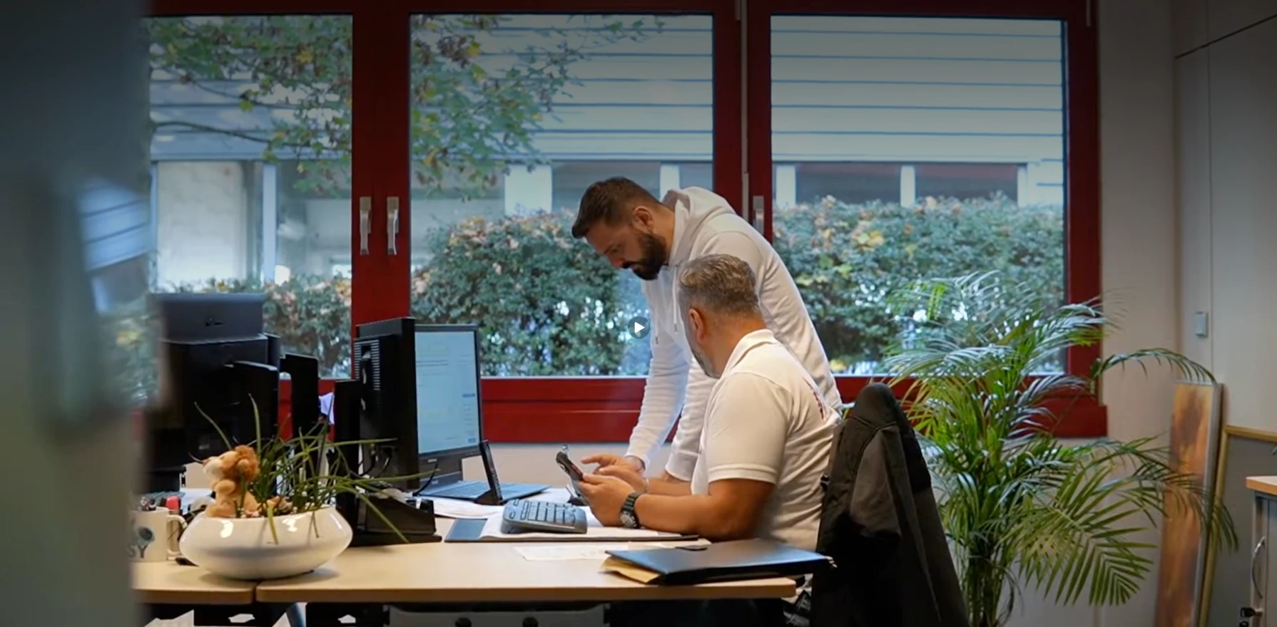 Zwei Männer arbeiten zusammen im Büro an einem Schreibtisch mit Computern und Pflanzen am Fenster.