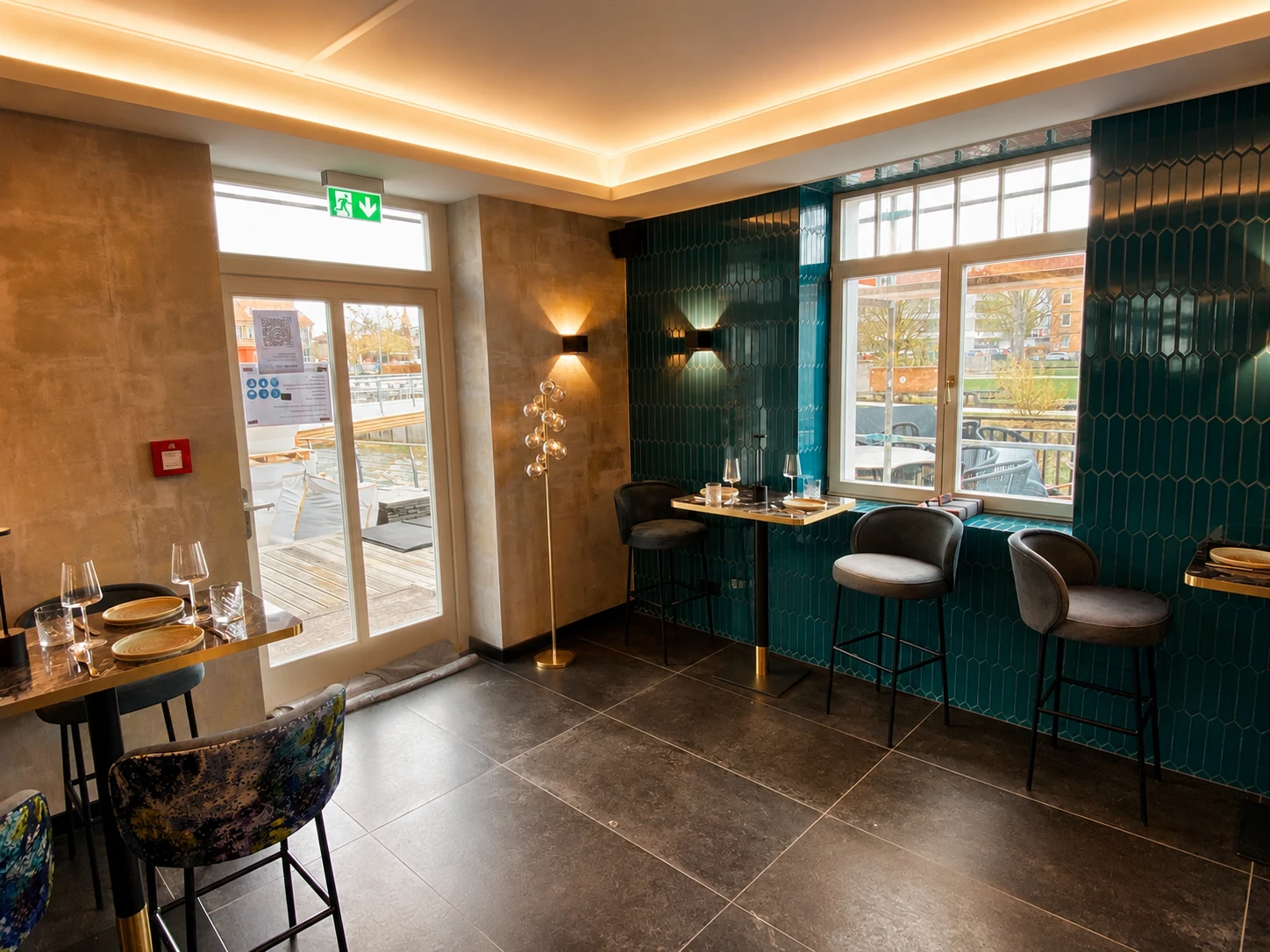 Modernes Café-Interieur mit dunkel gefliestem Boden, blauen Wandfliesen, Tischen mit Stühlen und großen Fenstern.