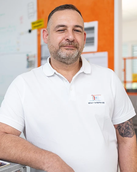 Mann mit kurzem Bart und Tattoo, der ein weißes Polohemd vor einem orangefarbenen Hintergrund trägt.