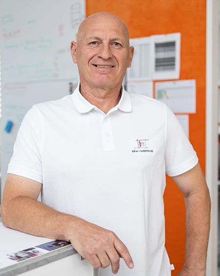 Älterer Mann in weißem Polohemd lehnt an Tisch vor einer Wand mit weißen und orangenen Bereichen.
