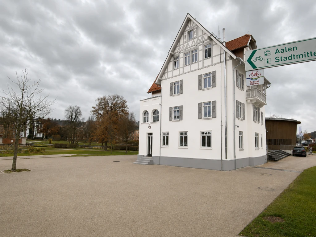 Weißes Fachwerkhaus neben Schildern, die auf Aalen und Stadtmitte hinweisen, unter bewölktem Himmel.