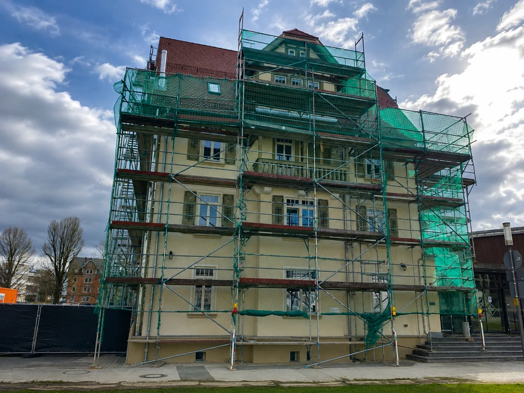 Haus mit grünen Gerüsten, das außen saniert wird, unter bewölktem Himmel.