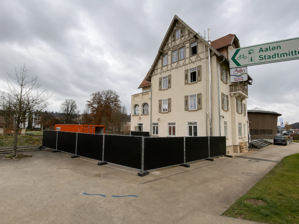 Traditionelles Fachwerkhaus neben Baustelle mit schwarzen Bauzäunen unter bewölktem Himmel.