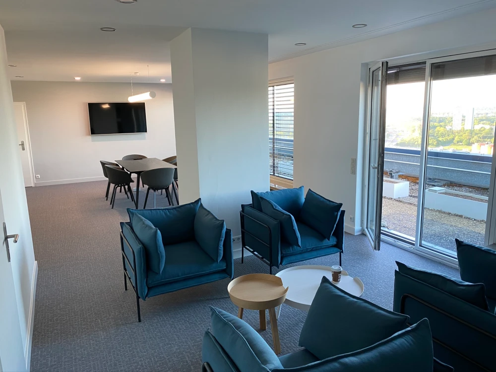 Modern office lounge with blue armchairs, round tables, dining area, and large window terrace door.