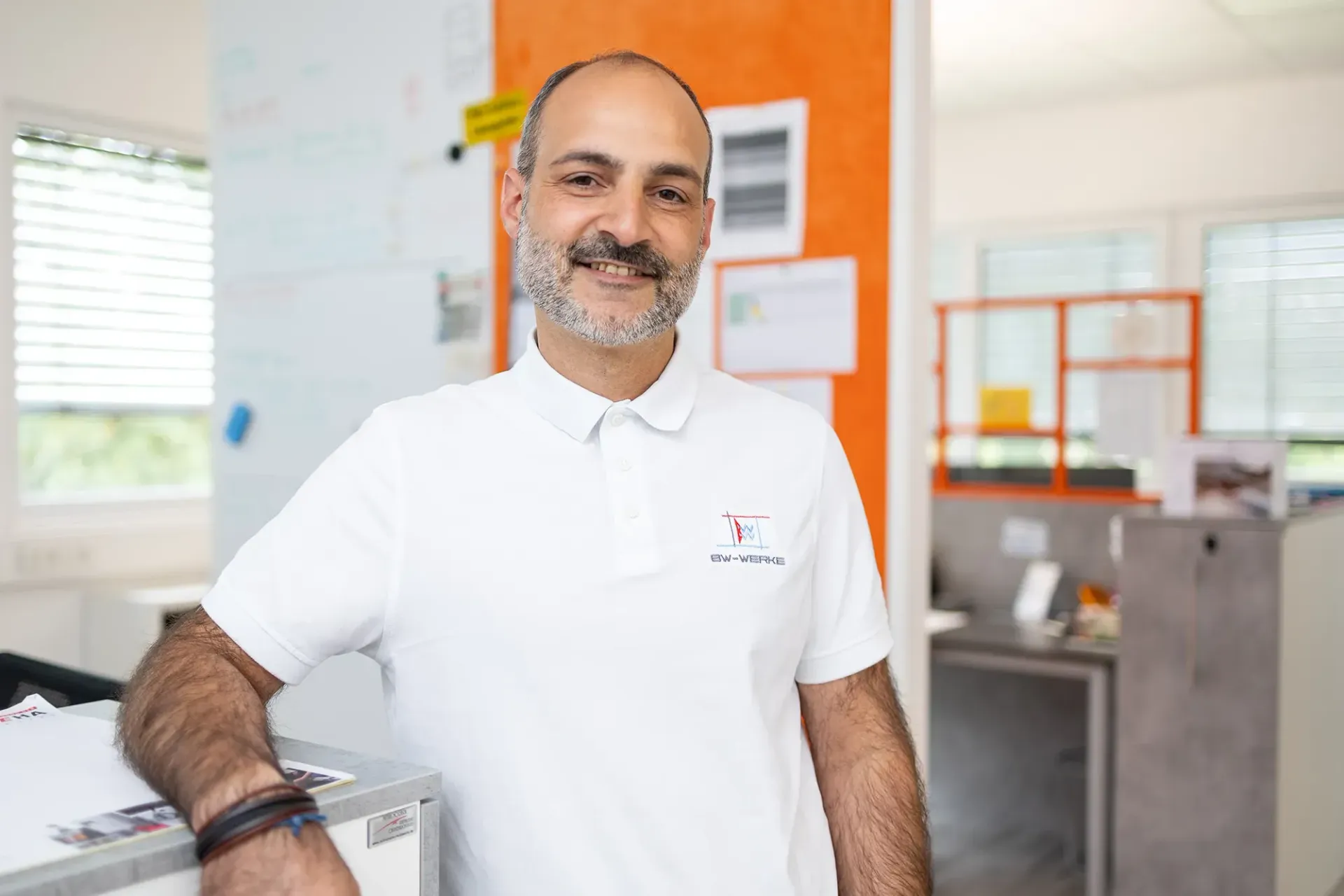 Ein lächelnder Mann mit grauem Bart trägt ein weißes Polohemd mit dem Logo BW-WERKE und lehnt sich in einem hellen Büro mit orangefarbenem Pinnwandbereich entspannt an eine Ablage.