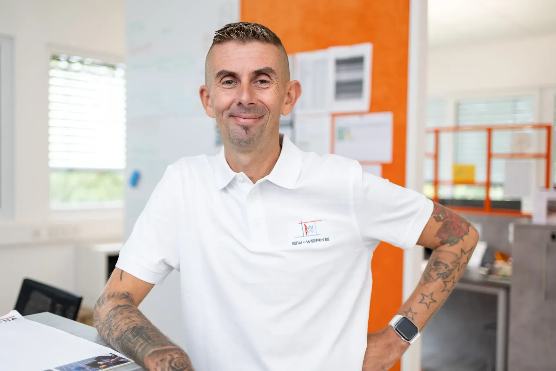 Mann mit kurzen Haaren und Tätowierungen an beiden Armen trägt ein weißes Poloshirt mit BW-Werke-Logo und steht lächelnd in einem hellen Büro mit orangefarbener Wand im Hintergrund.