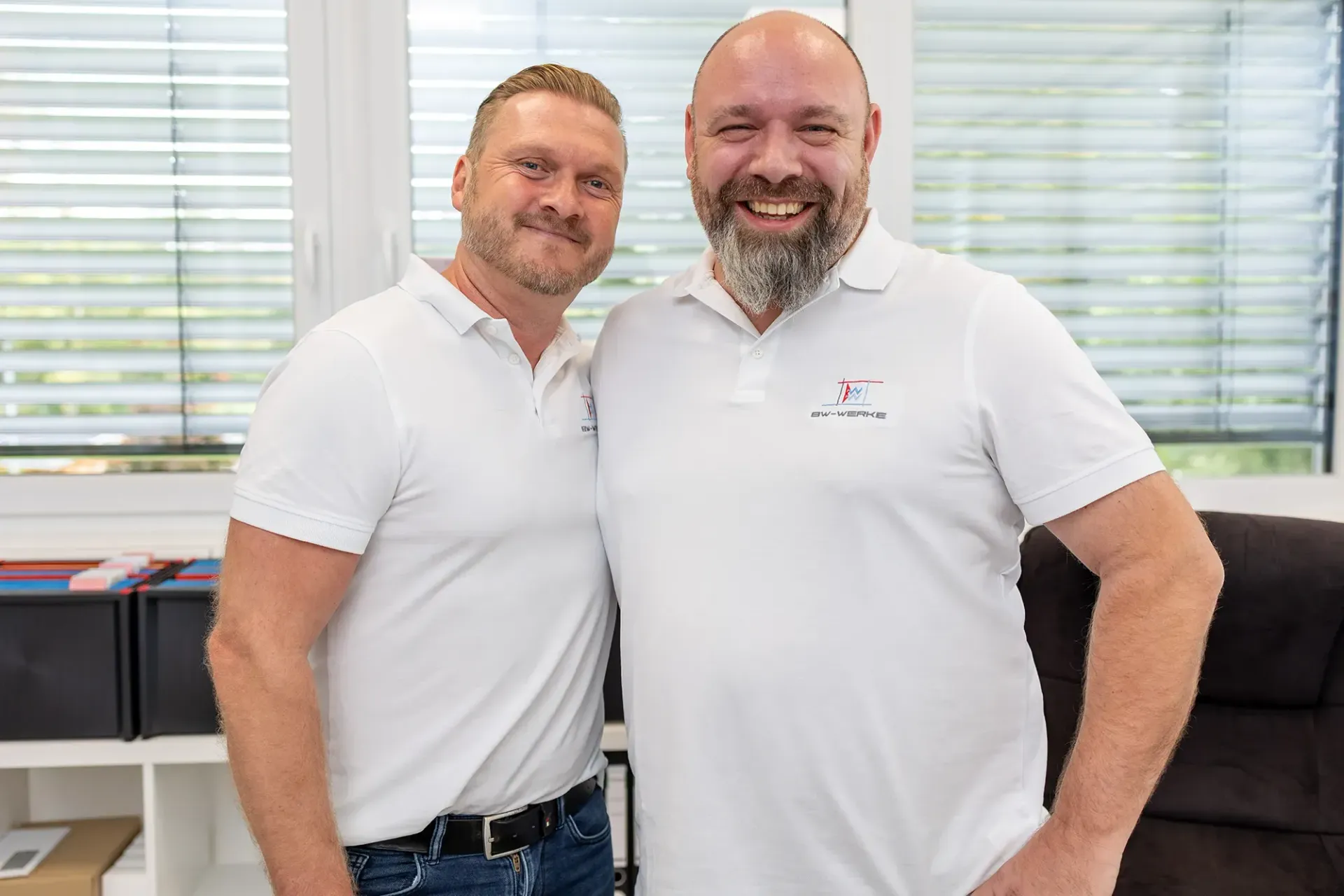 Zwei lachende Männer in weißen Poloshirts mit dem Logo BW-WERKE umarmen sich in einem hellen Büro mit Fensterjalousien.