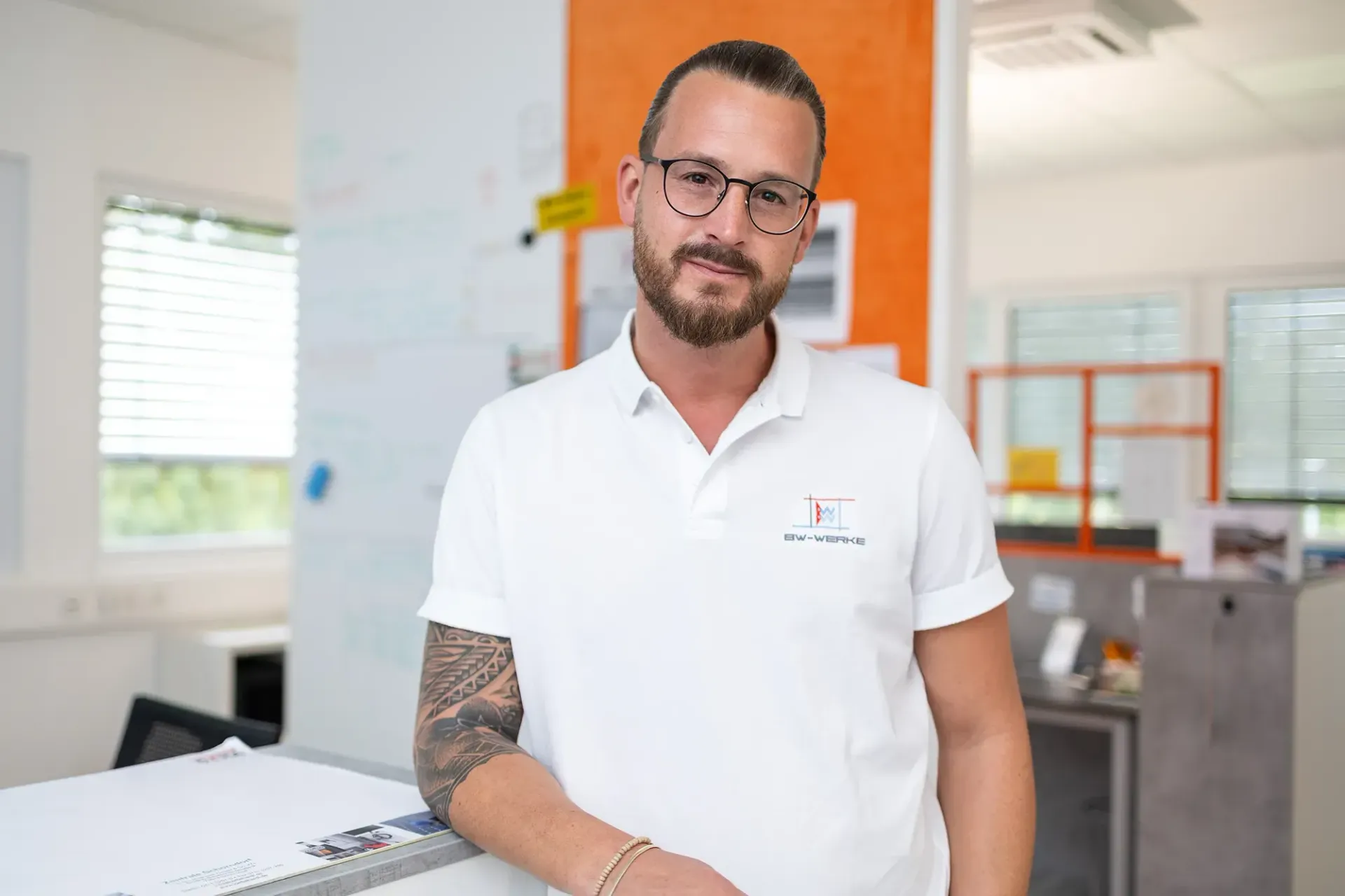 Mann mit Brille und Bart, trägt weißes Poloshirt mit BW-WERKE-Logo, mit tätowiertem rechten Arm, lehnt sich an weißen Tisch, Büro mit Fenstern und weißen Wänden im Hintergrund.