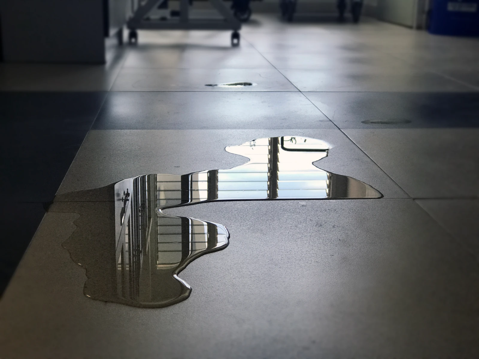 Wasserlache auf einem Fliesenboden, die das Fenster mit Gittern und Lichtreflexionen darüber spiegelt.