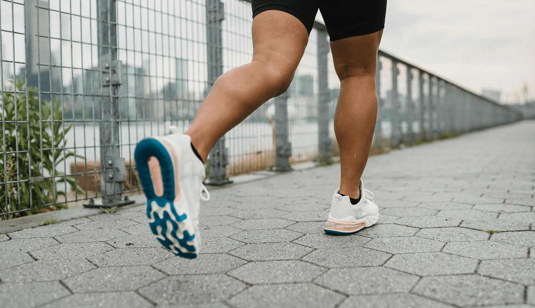 Close-up of a person jogging on a paved path near a wire fence, wearing white sneakers with blue soles. The scene conveys healthy movement and energy.