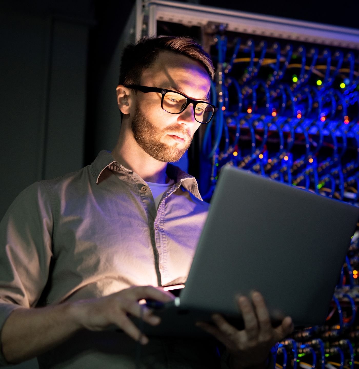 Man wearing glasses working on a laptop in a server room with illuminated network cables.