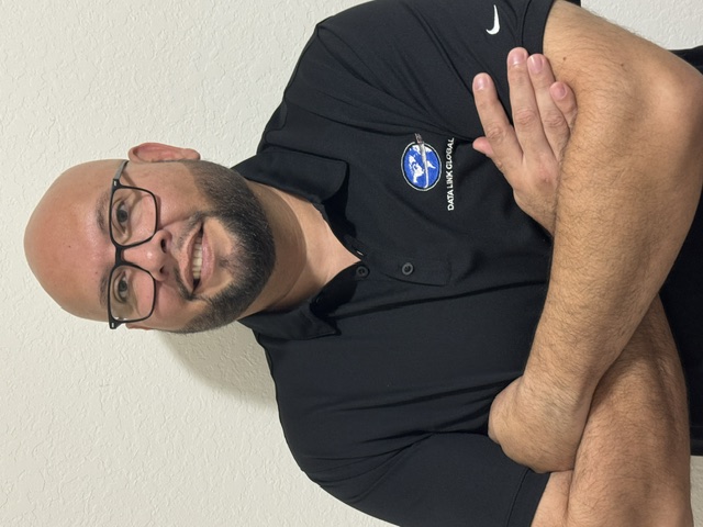 Smiling bald man with glasses wearing a black polo shirt with Data Link Global logo and a Nike swoosh, standing with arms crossed.