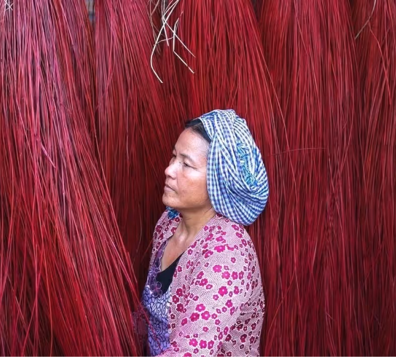 Woman wearing a patterned headscarf and floral jacket sitting among bundles of long red fibers hanging around her.