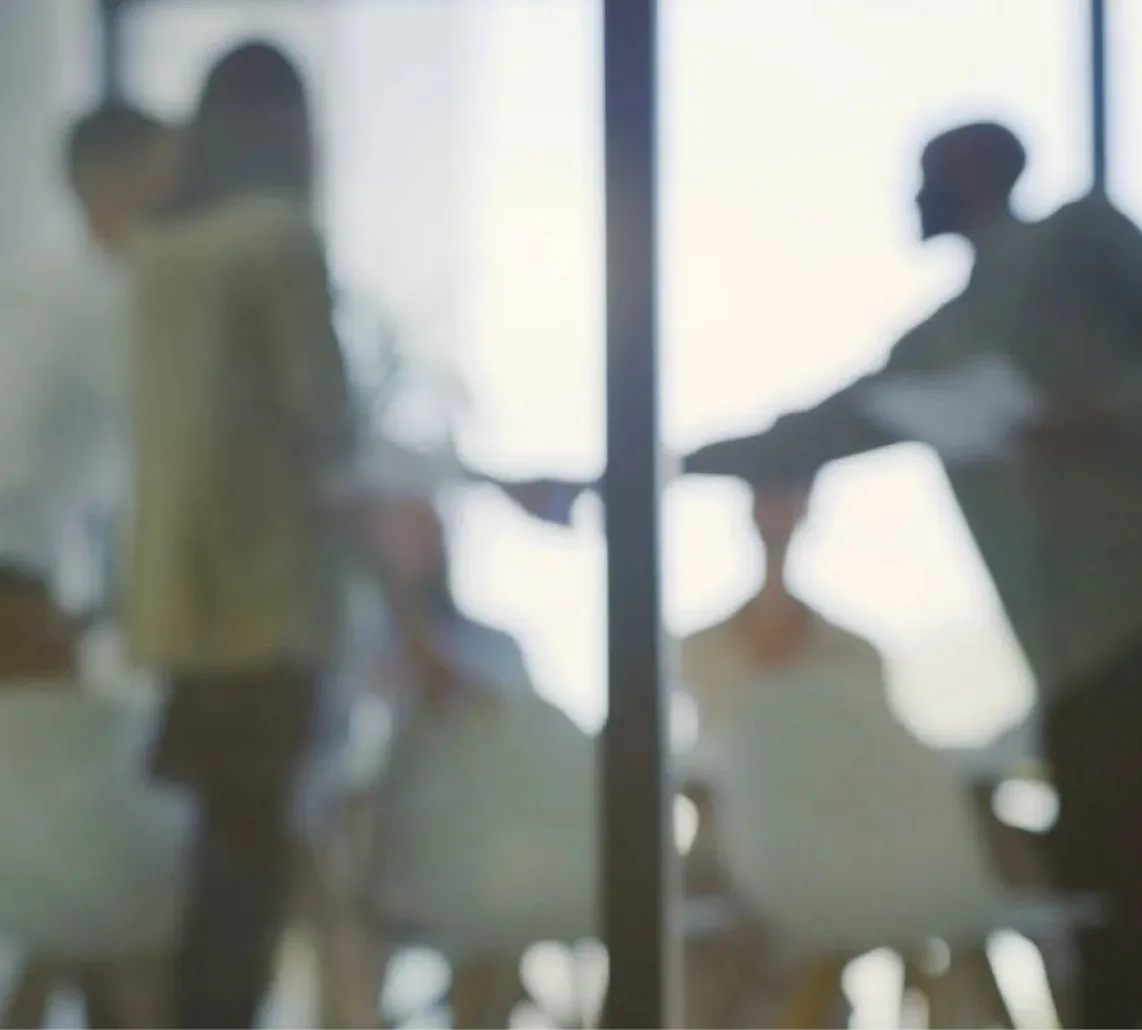 Blurry silhouettes of people in a meeting or discussion setting with bright windows in the background.