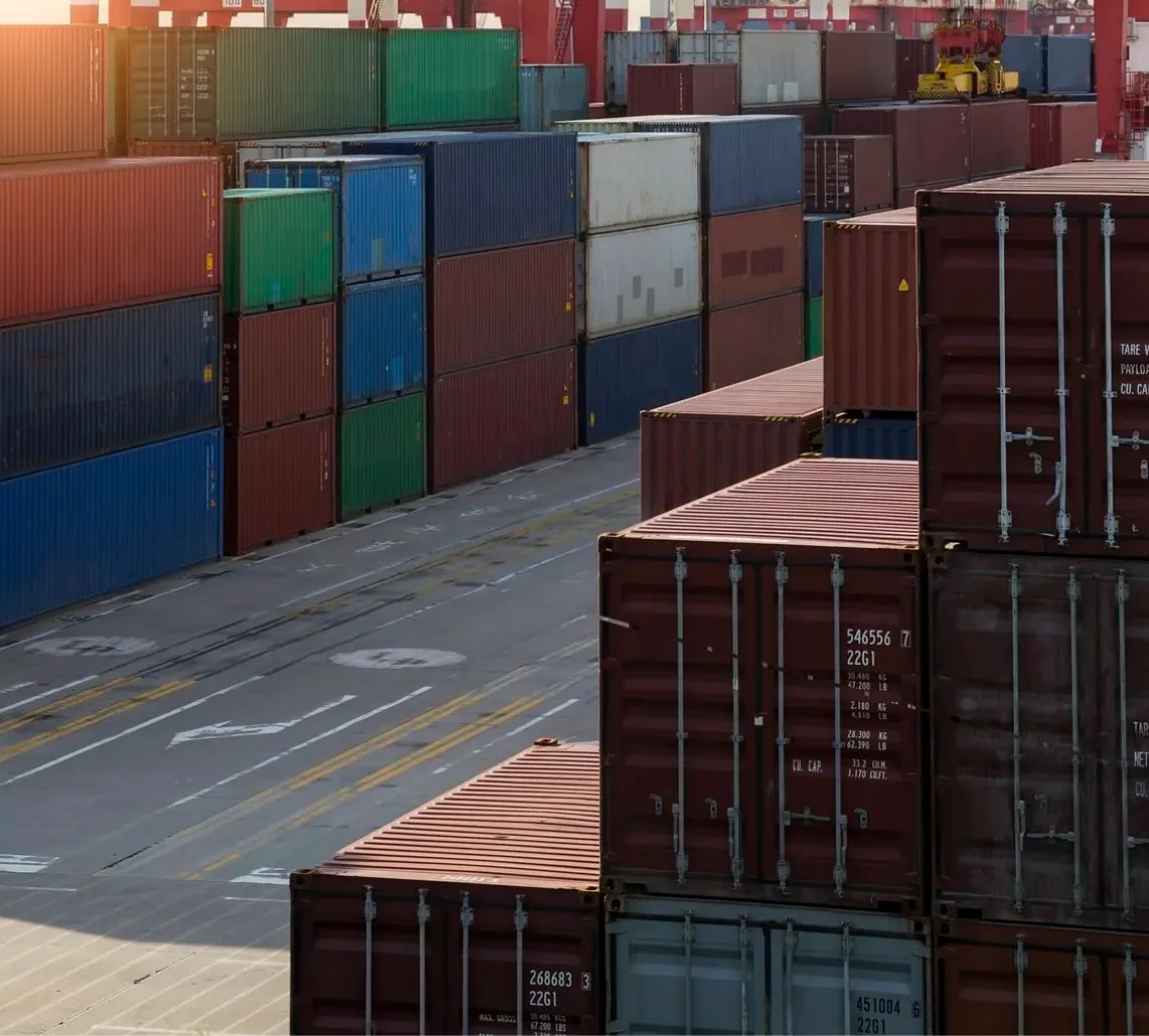 Stacks of multi-colored shipping containers arranged in rows at a cargo port during sunset.