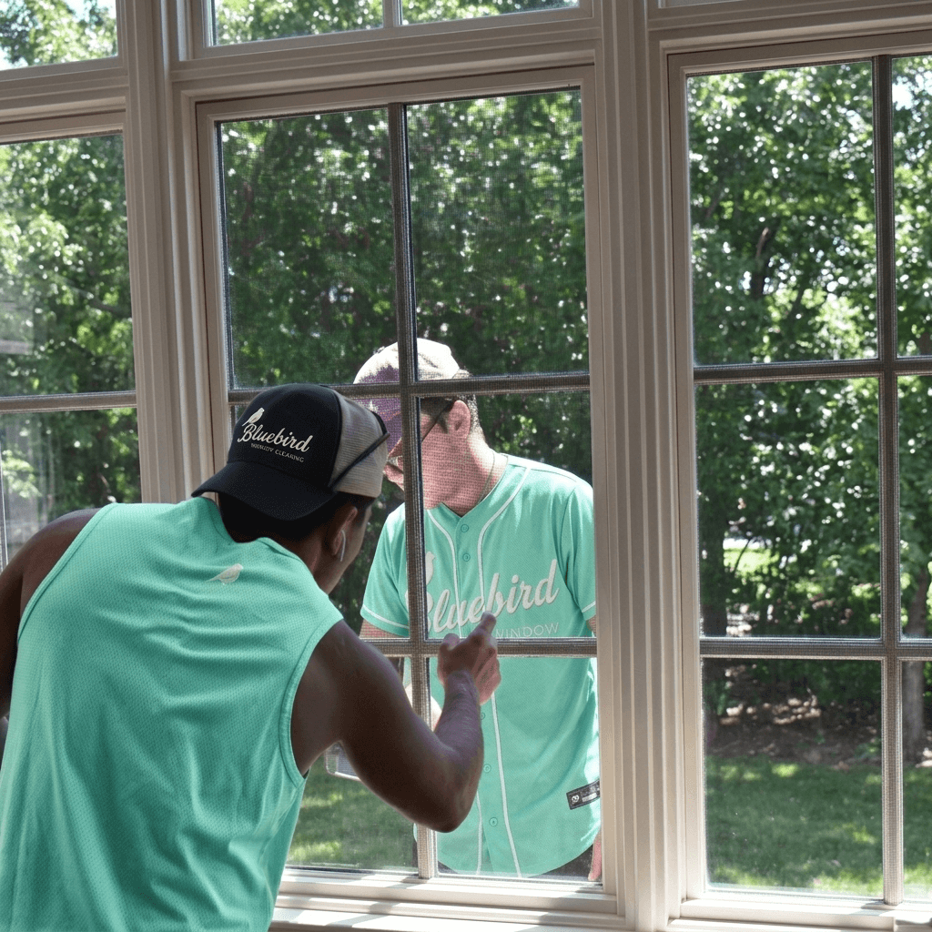 A man wearing a Bluebird Housekeeping Cleaning cap and turquoise shirt cleans the outside of a large window with a grid design.