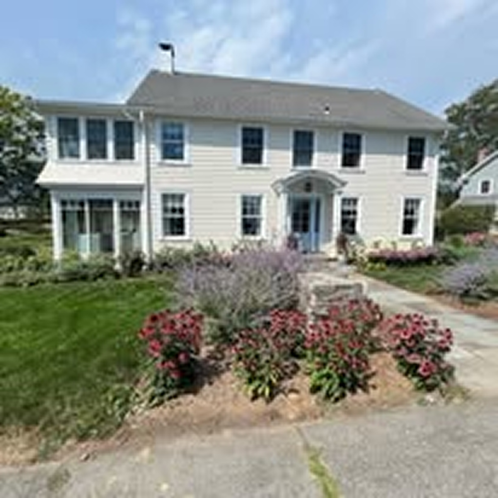 Two-story white house with multiple windows, front entrance with a small porch, surrounded by green lawn and flower beds.
