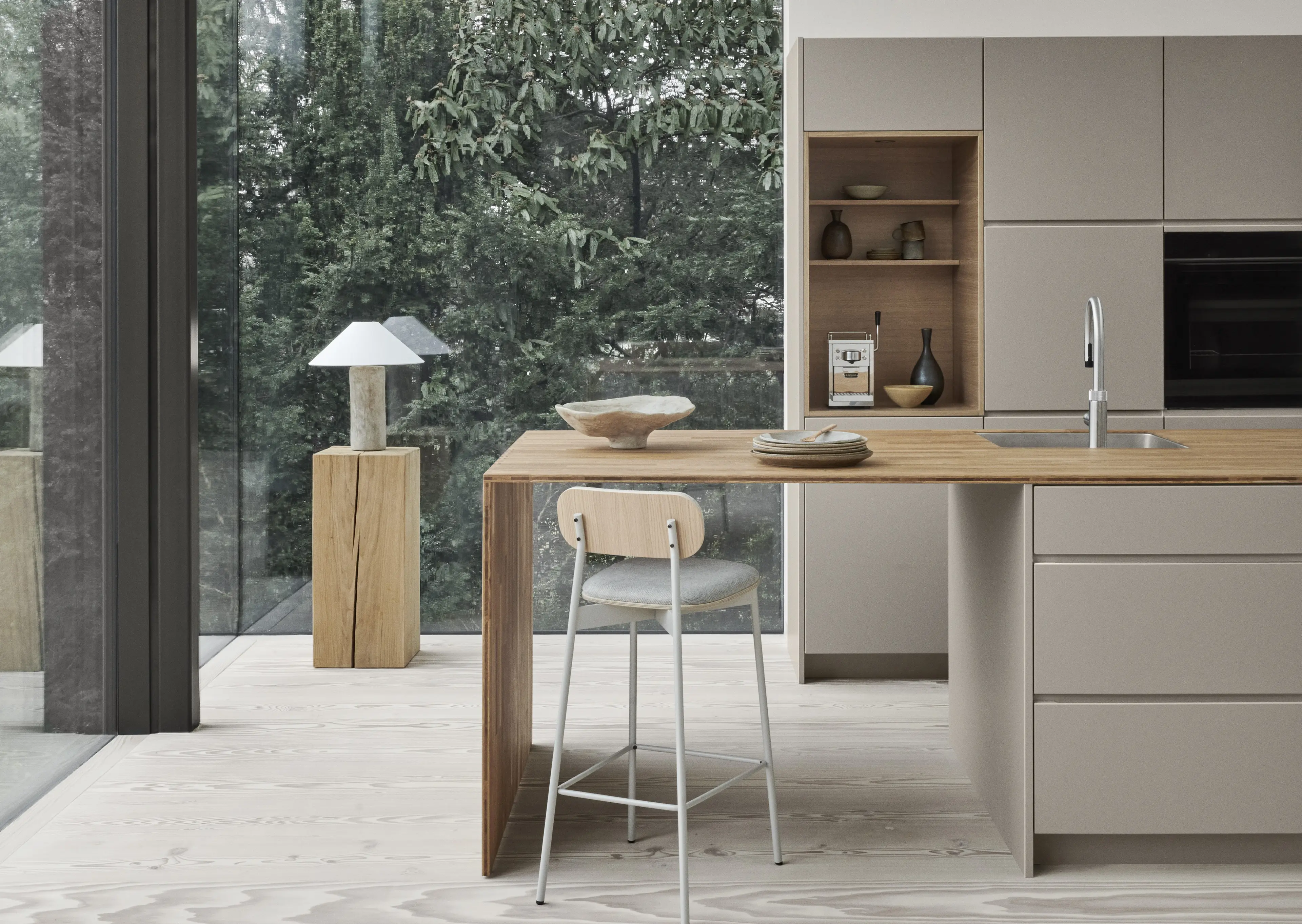 Modern kitchen with beige cabinetry, wooden island featuring a sink and countertop with plates and decorative bowl, and a stool with light wooden backrest and gray seat.