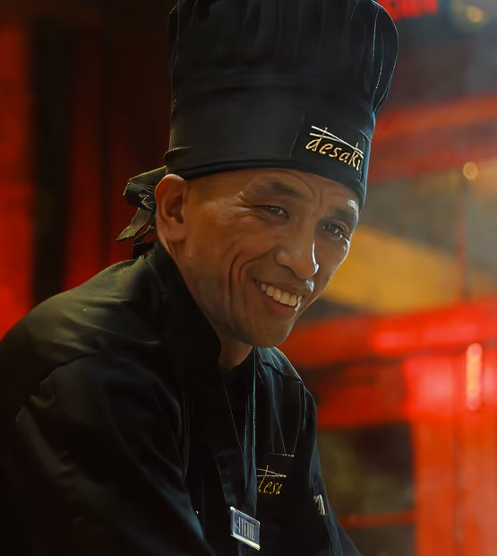 Smiling chef wearing a black uniform and tall black hat with 'tesaki' embroidered on them.