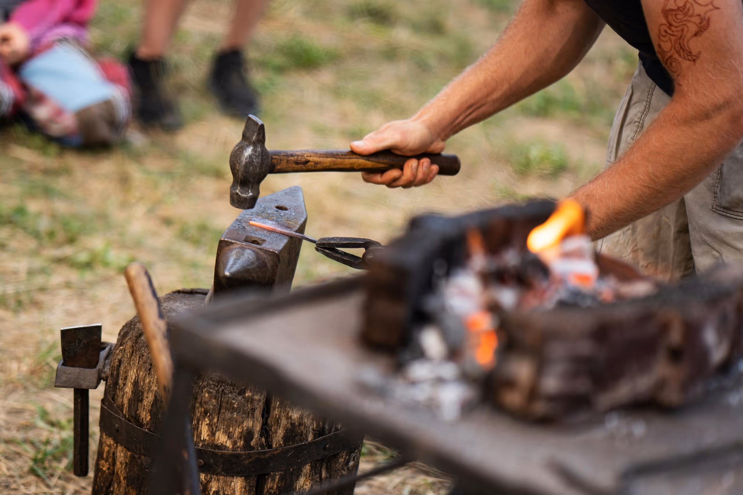Man hammering a heated metal rod on an anvil outdoors near a fire pit.