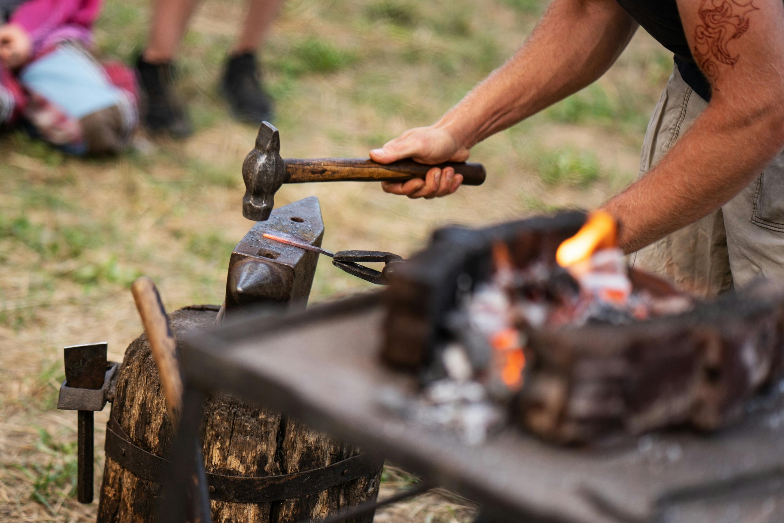 Man hammering a heated metal rod on an anvil outdoors near a fire pit.
