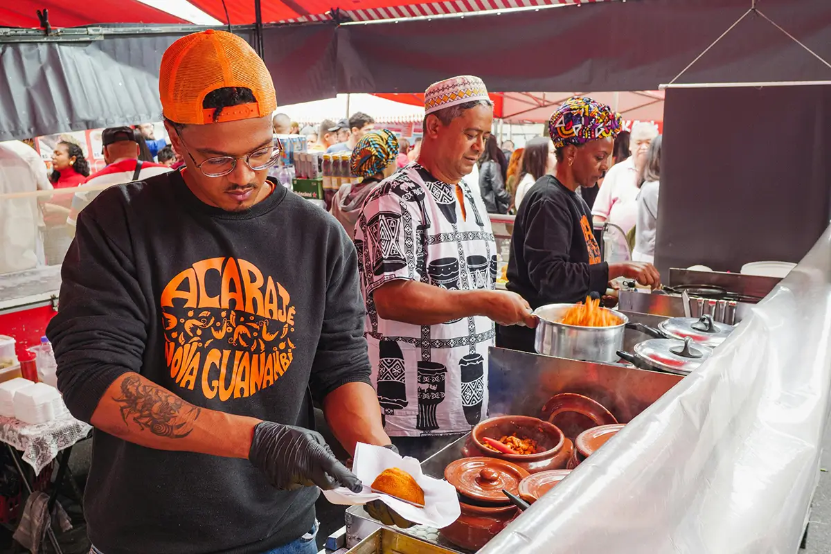 Three people working at a food stall preparing and serving traditional dishes, with one man holding a fried snack on a paper napkin.