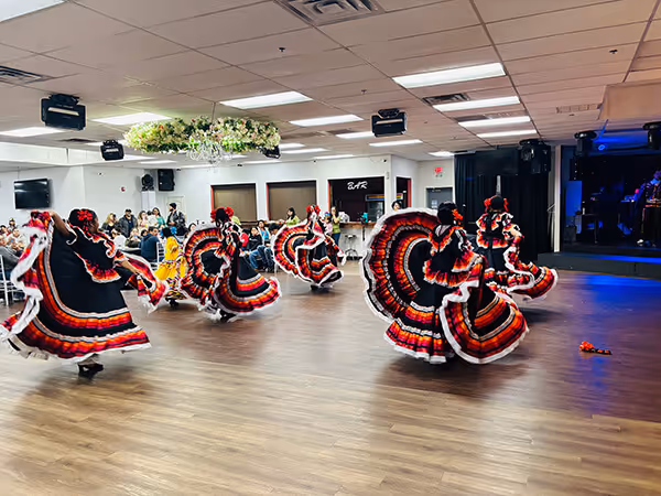 Group of dancers in black dresses with red, orange, and white trim performing a traditional dance on a wooden floor with an audience seated in the background.