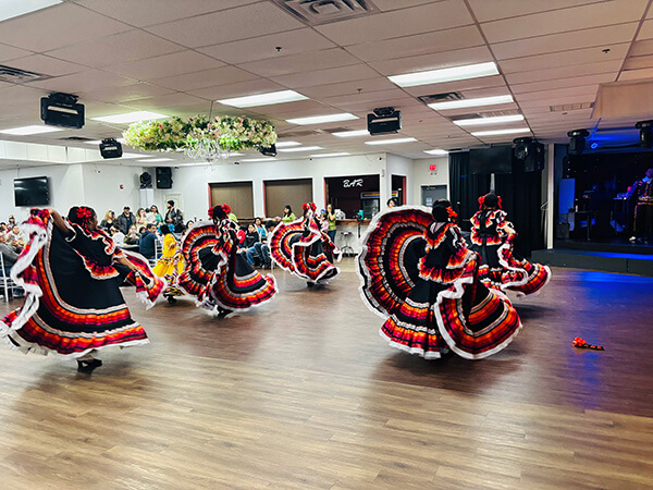 Group of dancers in black dresses with red, orange, and white trim performing a traditional dance on a wooden floor with an audience seated in the background.