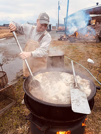 Man in overalls stirring a large steaming black pot over an outdoor fire with wood burning in the background.