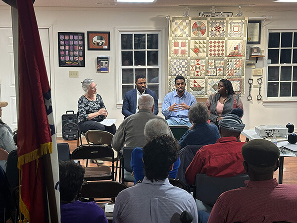 A discussion panel with four people seated facing an audience in a room with quilt and framed pictures on the wall.
