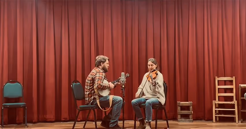 A man playing a banjo and a woman playing a violin seated on chairs on a wooden stage with a red curtain backdrop.