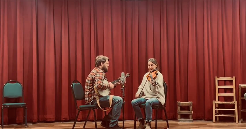 A man playing a banjo and a woman playing a violin seated on chairs on a wooden stage with a red curtain backdrop.