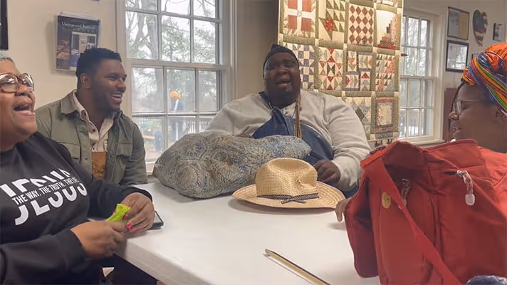 Four people sitting around a table indoors, smiling and singing together with a quilt hanging on the wall behind them.