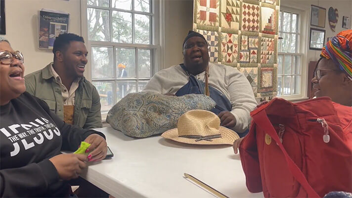 Four people sitting around a table indoors, smiling and singing together with a quilt hanging on the wall behind them.