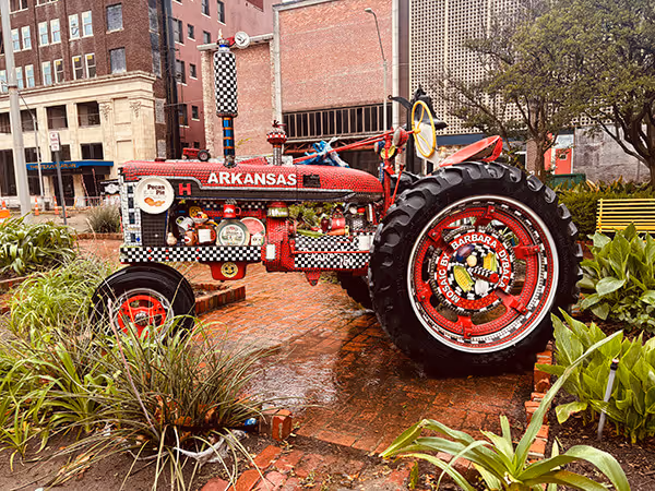 Red vintage Arkansas tractor displayed outdoors on a brick surface surrounded by plants and buildings.