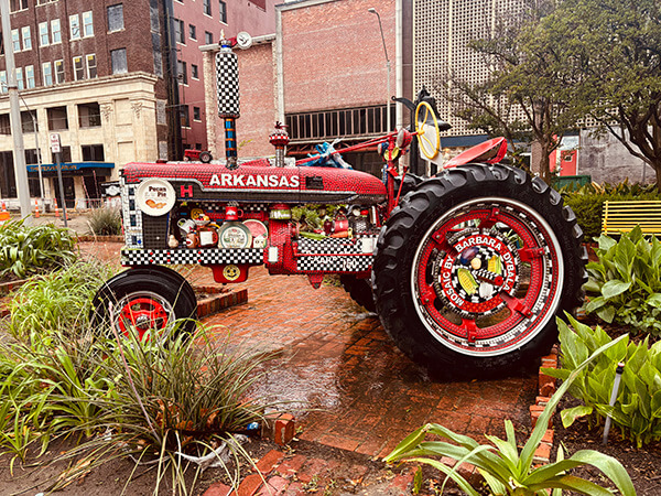 Red vintage Arkansas tractor displayed outdoors on a brick surface surrounded by plants and buildings.