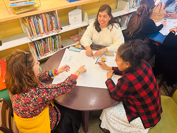 Three women seated around a round table writing and drawing on a large sheet of paper in a library setting.