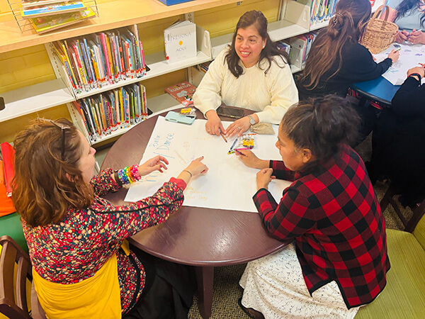 Three women seated around a round table writing and drawing on a large sheet of paper in a library setting.