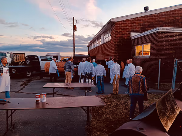Group of people in aprons gathered outside a brick building at dusk with picnic tables and a barbecue grill nearby.