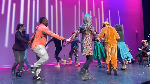 Group of people holding hands and square dancing on a stage with purple lighting.