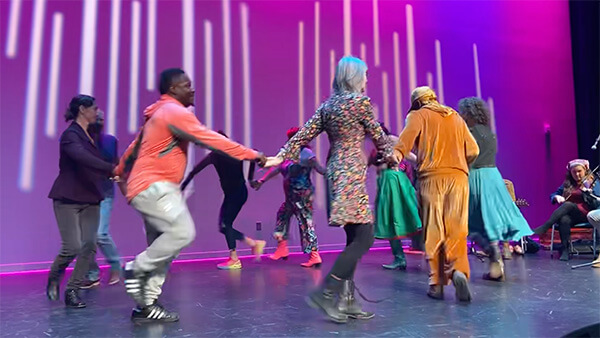 Group of people holding hands and square dancing on a stage with purple lighting.