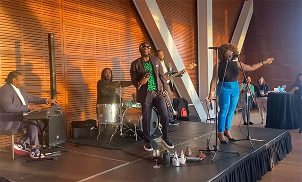 Band performing on stage with a keyboard player, drummer, bassist, male vocalist in sunglasses, and female vocalist in blue pants.