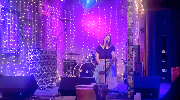 Musician sitting and playing a harp on a stage decorated with string lights and drums in the background.